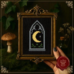 May include: Cross-stitch art in a gold ornate frame. The artwork features a black background with a yellow crescent moon, a white star, and green foliage. The frame is held by a hand with a red wax seal that reads "The Gilded Moth".