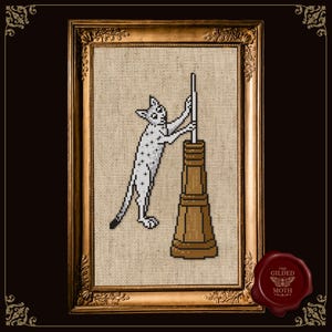 Puede incluir: Obra de arte de punto de cruz de un gato blanco con manchas negras, interactuando con una estructura marrón. La obra está enmarcada en un marco dorado adornado. El logotipo de The Gilded Moth está en la esquina inferior derecha.