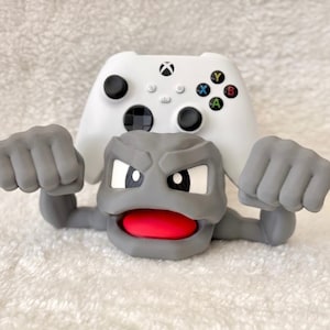 May include: A gray cartoon character with a red mouth and white eyes, holding up a white Xbox controller. The controller has black buttons and colorful A, B, X, and Y buttons.