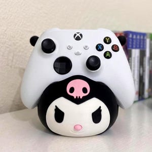Puede incluir: Un mando Xbox blanco descansa sobre una base en blanco y negro con temática de personaje. El mando tiene joysticks negros y botones de colores. La base tiene un diseño de calavera y huesos cruzados con una nariz rosa.