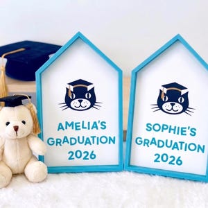 May include: Two house-shaped graduation frames with blue trim, each displaying a cat wearing a graduation cap and the text "AMELIA'S GRADUATION 2026" and "SOPHIE'S GRADUATION 2026". A teddy bear in a graduation cap and a mortarboard are also present.