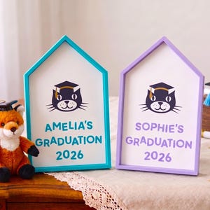 May include: Two house-shaped graduation signs, one teal and one purple, with a cat wearing a graduation cap. The teal sign reads "Amelia's Graduation 2026," and the purple sign reads "Sophie's Graduation 2026." A stuffed fox wearing a graduation cap sits nearby.