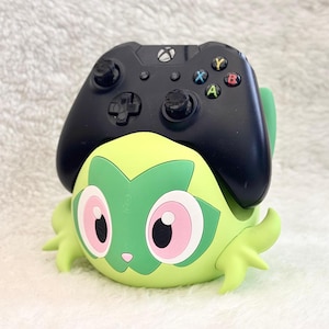 May include: A black Xbox wireless controller resting on a light green, stylised character stand. The stand features large pink eyes, a dark green facial marking, and small pointed limbs, designed to hold a gaming controller securely in place.