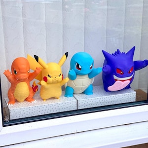 May include: Four colourful plastic character figurines standing in a row. From left to right, an orange lizard-like creature with a flame tail, a yellow mouse-like creature with long ears, a blue turtle-like creature, and a purple round creature with red eyes.