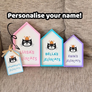 May include: Three house-shaped wooden signs with pink, blue, and purple frames, and one rectangular tag with a ribbon. Each features a black cat illustration with an orange bell on its head and the text "Personalise your name!" at the top.