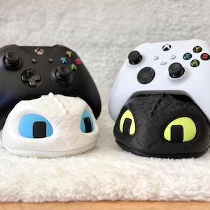 Puede incluir: Dos mandos de Xbox, uno negro y otro blanco, descansan sobre soportes con temática de dragón. El soporte blanco tiene ojos azules, mientras que el soporte negro tiene ojos amarillos. Los mandos tienen botones de colores y crucetas direccionales.