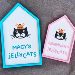May include: Two house-shaped signs with white backgrounds and colorful frames. Each sign features a black cat illustration wearing an orange hat. One sign reads "MACY'S JELLYCATS" in blue, the other reads "SAVANNAH'S JELLYCATS" in pink.