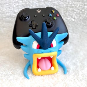 May include: A black Xbox wireless controller resting on a blue, yellow, and red character-shaped stand. The stand features a blue head with a dark blue crest, red eyes, a yellow mouth, and a pink tongue, designed to hold a gaming controller.