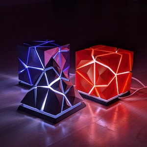 Könnte beinhalten: Drei geometrische, würfelförmige Lampen mit modernem Design. Eine Lampe strahlt blaues Licht aus, eine andere rotes, und die dritte eine Kombination aus beidem. Die Lampen haben eine facettierte Oberfläche und stehen auf einem schwarzen Sockel.