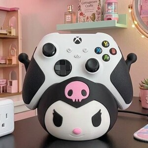 May include: A white Xbox controller sits atop a black and white Kuromi-themed base. The controller features black thumbsticks and coloured buttons. A pink skull is centered on the base. Shelves with plush toys and a dollhouse are in the background.