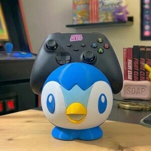K&ouml;nnte beinhalten: Ein blau-wei&szlig;-gelber Piplup-Controllerhalter mit einem schwarzen Xbox-Controller obenauf. Der Halter steht auf einer Holzoberfl&auml;che, mit einem Retro-Arcade-Spiel und anderem Gaming-Zubeh&ouml;r im Hintergrund.
