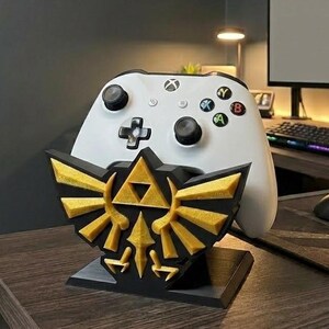 May include: A white Xbox controller sits on a black and gold decorative stand. The stand features a gold emblem with a black base. The controller has black buttons and coloured action buttons.