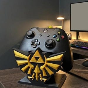 May include: A black Xbox controller rests on a custom-made stand featuring the gold and black emblem of the Legend of Zelda. The stand is on a wooden desk, with a computer monitor and keyboard in the background.