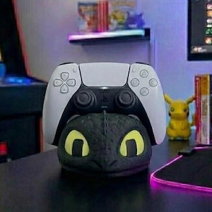 May include: A white and black PlayStation 5 controller sits atop a black dragon-shaped stand with yellow eyes. A yellow Pikachu figure and game cases are in the background. The scene is set on a black desk.