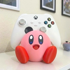 May include: A pink Kirby figurine with red feet and a smiling expression is positioned in front of a white Xbox controller. The controller has black buttons and colored action buttons. A second controller is visible in the background.