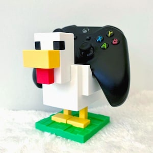 Puede incluir: Soporte para mando con forma de gallina blanca, amarilla, roja y negra, sosteniendo un mando Xbox negro. La base verde simula hierba, creando un diseño lúdico y original.