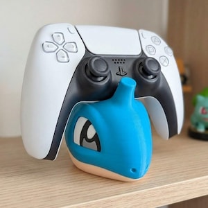 May include: A white and black PlayStation 5 controller rests above a blue Lapras-themed controller stand. The stand is light blue with a tan base and features Lapras's face. The controller has black joysticks and buttons.