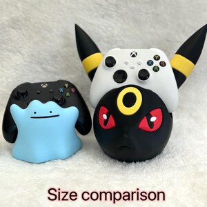 May include: A light blue Ditto-shaped controller stand next to a black Xbox controller, and a black and yellow Umbreon-shaped controller stand holding a white Xbox controller. The text "Size comparison" is visible at the bottom of the image.