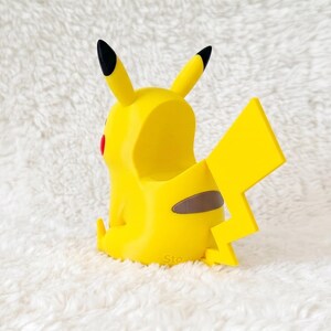 May include: A yellow Pikachu-shaped figurine with a hollow back, featuring two pointed ears with black tips, a jagged lightning bolt-shaped tail, and a small brown patch on the lower back. The character has a red circular cheek visible on the side.
