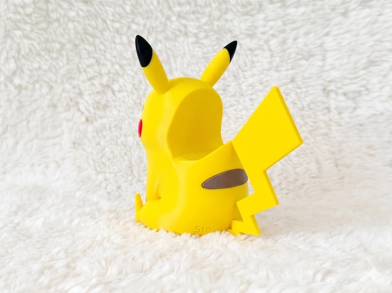 May include: A yellow Pikachu-shaped figurine with a hollow back, featuring two pointed ears with black tips, a jagged lightning bolt-shaped tail, and a small brown patch on the lower back. The character has a red circular cheek visible on the side.
