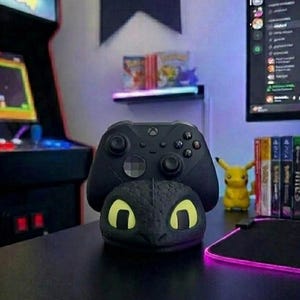 May include: A black Xbox controller sits on a black dragon head stand with yellow eyes. The stand is on a black desk. An arcade machine and computer monitor are in the background. A Pikachu figure and game cases are also visible.