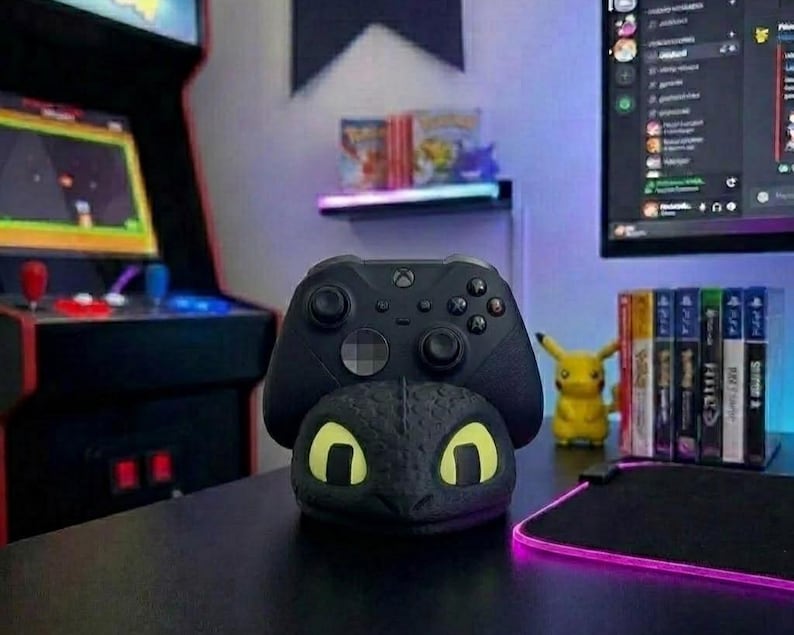 May include: A black Xbox controller sits on a black dragon head stand with yellow eyes. The stand is on a black desk. An arcade machine and computer monitor are in the background. A Pikachu figure and game cases are also visible.
