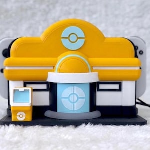 May include: A yellow and white plastic gaming console dock shaped like a building with a light blue circular emblem on the front. The structure features a central door, a small side kiosk, and a dark base, designed to hold a handheld gaming device.