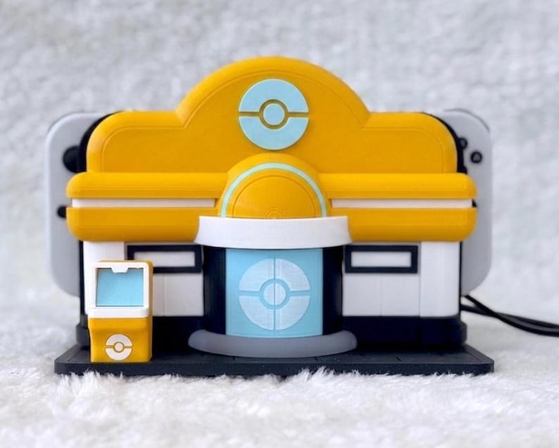 May include: A yellow and white plastic gaming console dock shaped like a building with a light blue circular emblem on the front. The structure features a central door, a small side kiosk, and a dark base, designed to hold a handheld gaming device.