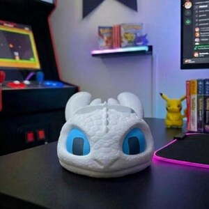 May include: A white dragon-shaped desk tidy with blue eyes rests on a black desk. The organiser is in front of a gaming setup, including a monitor, arcade machine, and a Pikachu figurine.