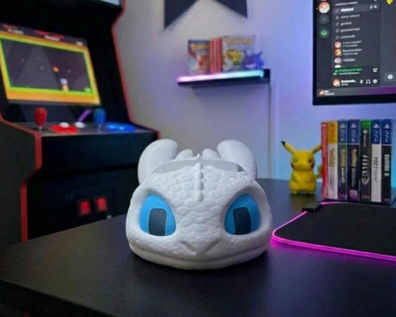 May include: A white dragon-shaped desk tidy with blue eyes rests on a black desk. The organiser is in front of a gaming setup, including a monitor, arcade machine, and a Pikachu figurine.