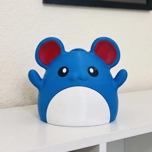 May include: A blue and white figurine with red ears and black eyes. The figurine has a round body with small arms and is sitting on a white surface. The figurine is a cartoon character.