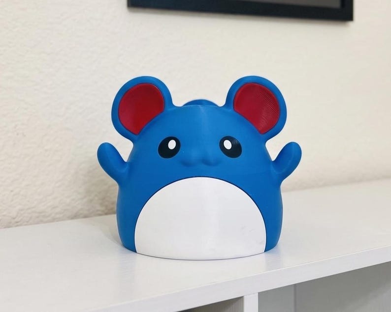 May include: A blue and white figurine with red ears and black eyes. The figurine has a round body with small arms and is sitting on a white surface. The figurine is a cartoon character.