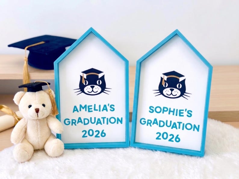 May include: Two house-shaped graduation frames with blue trim, each displaying a cat wearing a graduation cap and the text "AMELIA'S GRADUATION 2026" and "SOPHIE'S GRADUATION 2026". A teddy bear in a graduation cap and a mortarboard are also present.