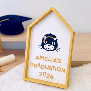 May include: A yellow framed graduation sign with the text "AMELIA'S GRADUATION 2026" and a cat wearing a graduation cap. A navy blue graduation cap and a rolled diploma are in the background.