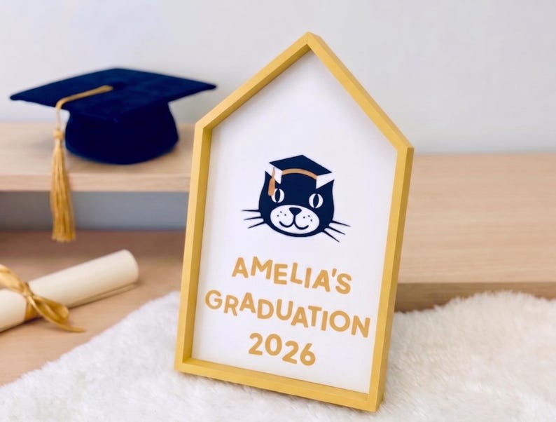 May include: A yellow framed graduation sign with the text "AMELIA'S GRADUATION 2026" and a cat wearing a graduation cap. A navy blue graduation cap and a rolled diploma are in the background.