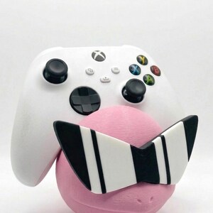 Puede incluir: Un mando blanco de Xbox descansa contra un objeto rosa y redondo que lleva gafas de sol a rayas blancas y negras. El mando tiene joysticks negros y botones de colores. El fondo es blanco liso.