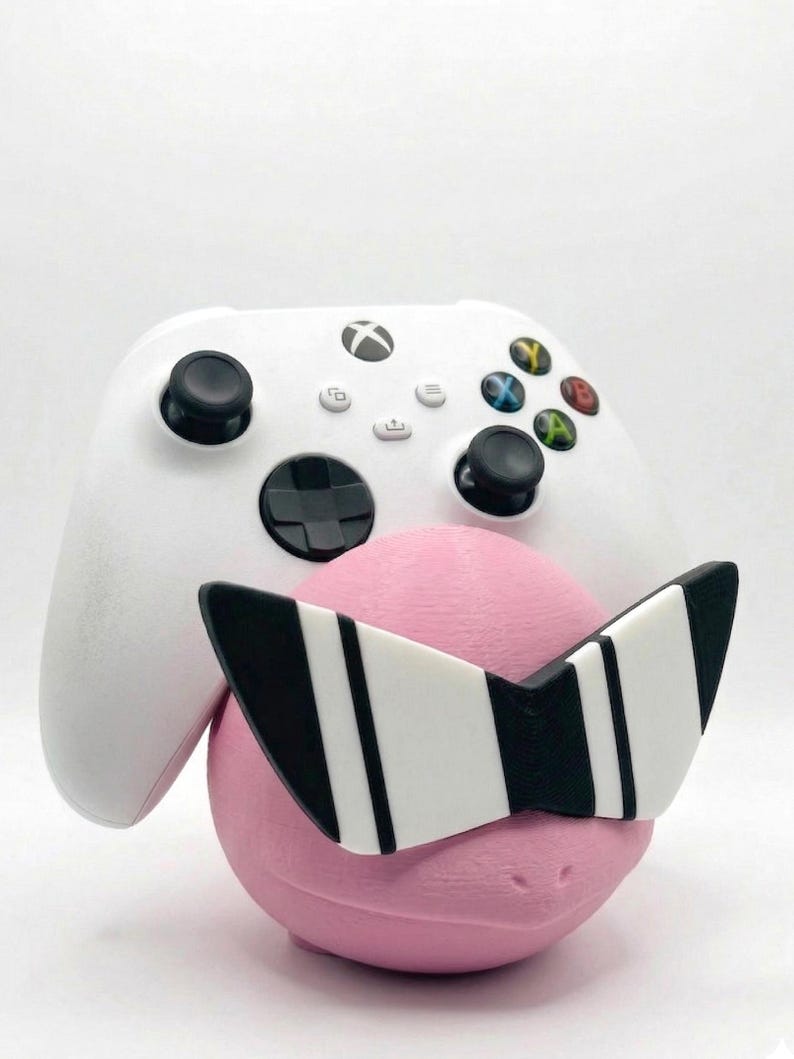 Puede incluir: Un mando blanco de Xbox descansa contra un objeto rosa y redondo que lleva gafas de sol a rayas blancas y negras. El mando tiene joysticks negros y botones de colores. El fondo es blanco liso.