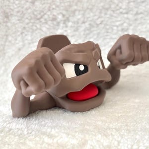 May include: A brown cartoon figure with a red mouth, white and black eyes, and raised fists. The figure is lying down on a white, textured surface. The figure's design is reminiscent of a popular animated character.