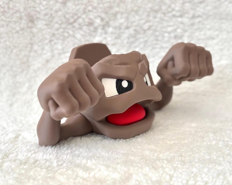 May include: A brown cartoon figure with a red mouth, white and black eyes, and raised fists. The figure is lying down on a white, textured surface. The figure's design is reminiscent of a popular animated character.