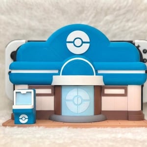 May include: A blue, white, and brown 3D-printed model of a building with a circular logo on the roof. The model has a light blue facade and a white and brown base. A white and black game console is behind the model.