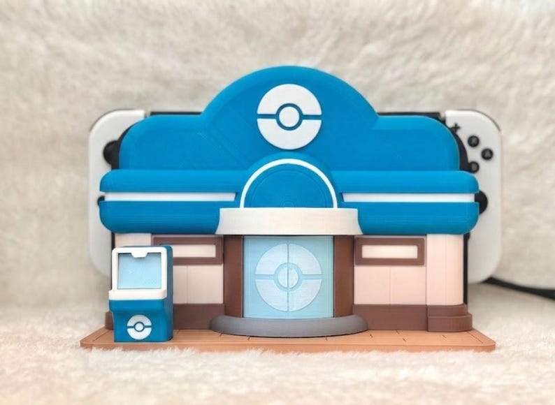 May include: A blue, white, and brown 3D-printed model of a building with a circular logo on the roof. The model has a light blue facade and a white and brown base. A white and black game console is behind the model.