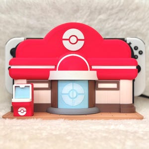 May include: A red and white building-shaped accessory with a circular logo on the roof and a blue and white circular design on the front. The accessory is placed in front of a white and black game console.