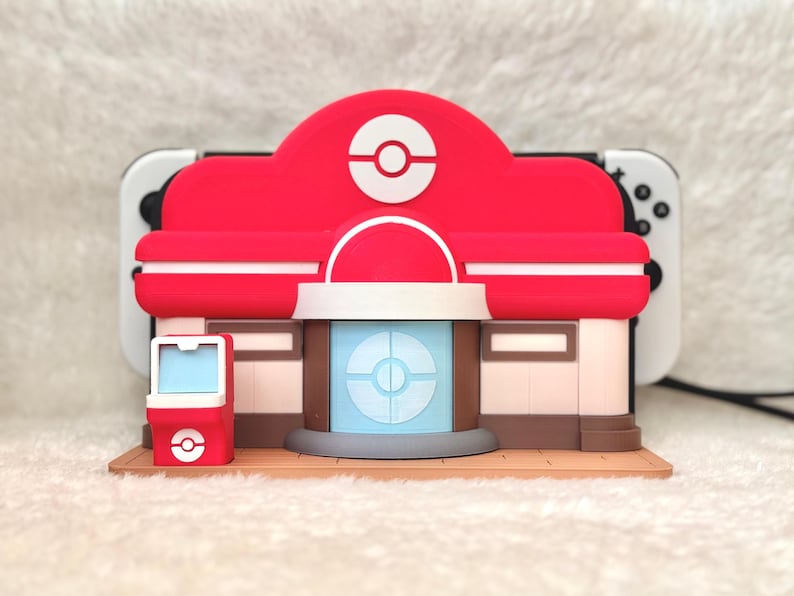 May include: A red and white building-shaped accessory with a circular logo on the roof and a blue and white circular design on the front. The accessory is placed in front of a white and black game console.