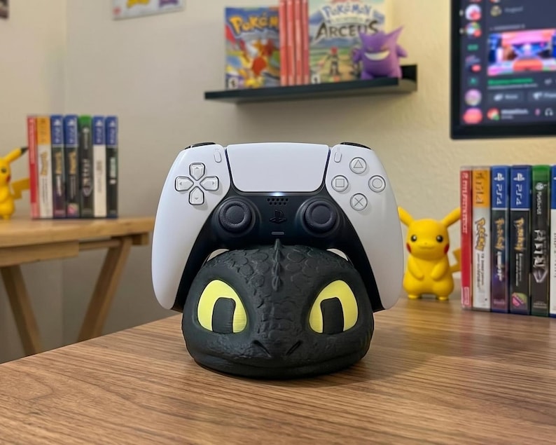 May include: A black dragon-themed controller stand supports a white and black PlayStation 5 controller. The stand features yellow eyes and a detailed dragon face. The controller is white with black buttons.