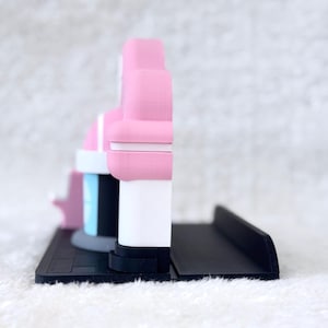 May include: A 3D printed display stand featuring a pink and white structure with a light blue accent, mounted on a black rectangular base. The object has a layered, matte finish with a distinct side profile showing a vertical design.