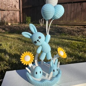 May include: A light blue knitted Easter basket with a bunny holding three balloon-shaped decorations. The basket contains two small bunnies, two yellow and white flowers, and a blue egg. The background includes a wooden fence and grass.