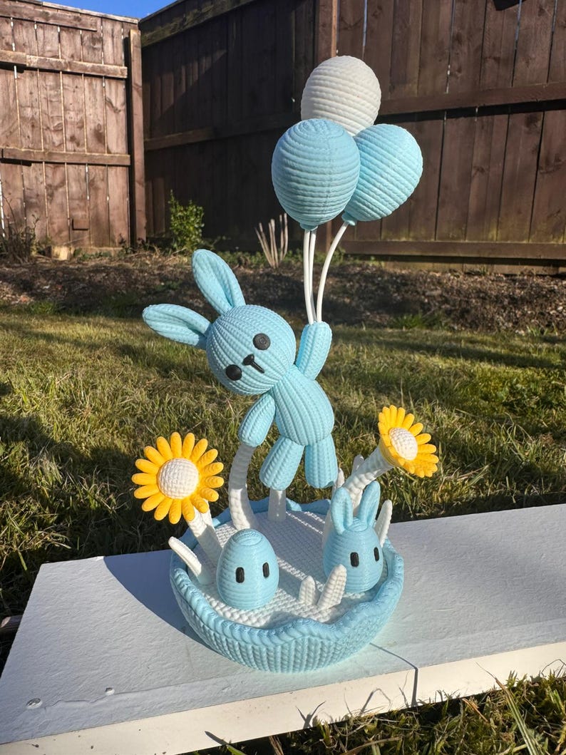 May include: A light blue knitted Easter basket with a bunny holding three balloon-shaped decorations. The basket contains two small bunnies, two yellow and white flowers, and a blue egg. The background includes a wooden fence and grass.