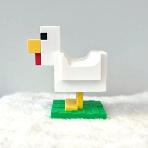 May include: A white, blocky chicken figurine with a yellow beak and legs, a red wattle, and a black eye. The chicken stands on a green base with a grid pattern. The figurine is likely a decorative item or toy.
