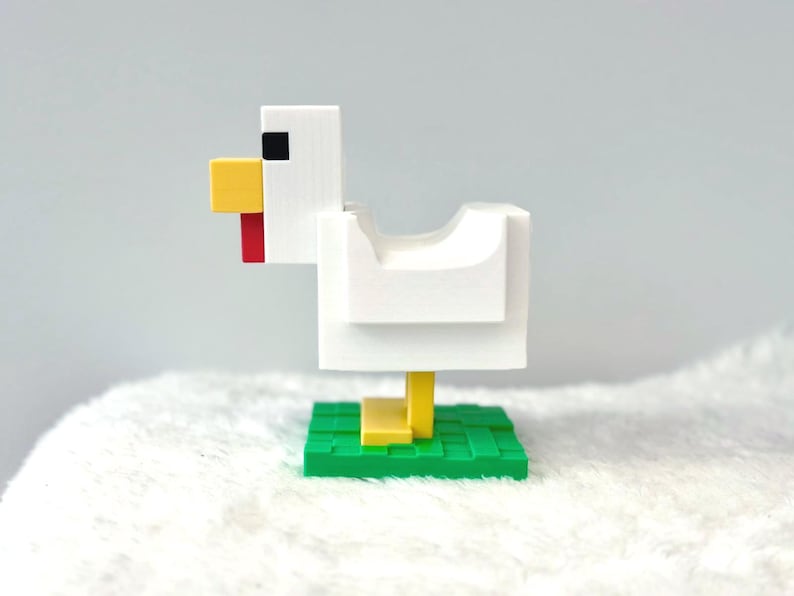 May include: A white, blocky chicken figurine with a yellow beak and legs, a red wattle, and a black eye. The chicken stands on a green base with a grid pattern. The figurine is likely a decorative item or toy.