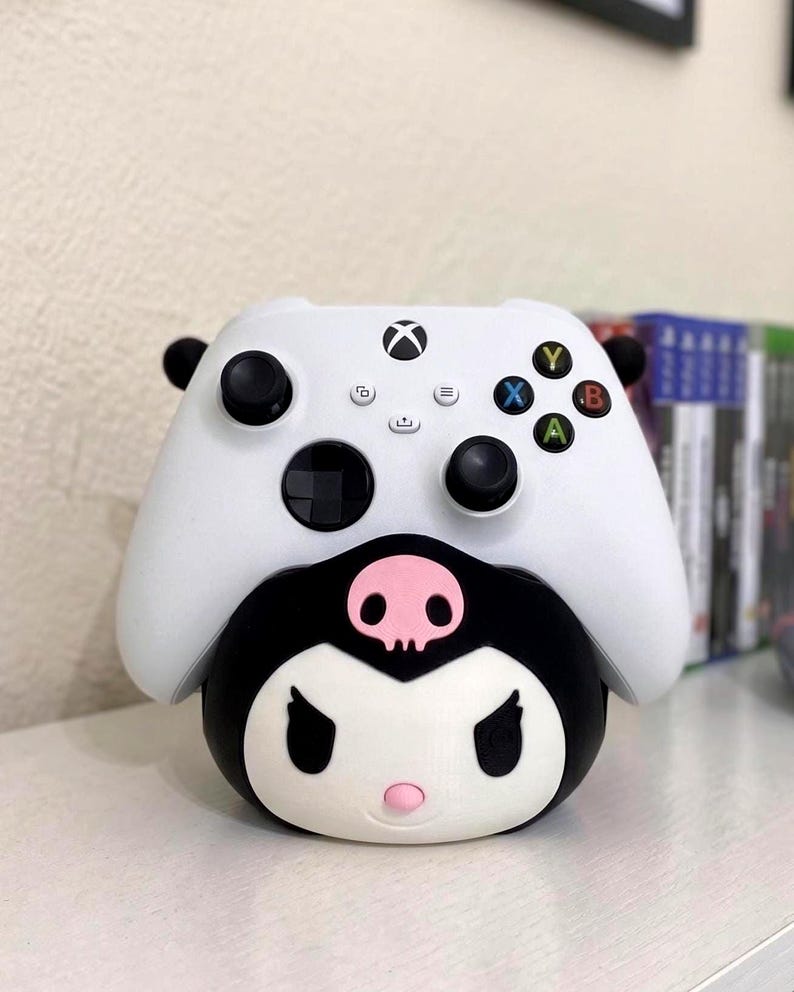 May include: A white Xbox wireless controller resting on a black and white character-themed stand. The stand features a stylized face with a pink skull emblem on the forehead, black ears, and a small pink nose, designed to hold the gaming controller.